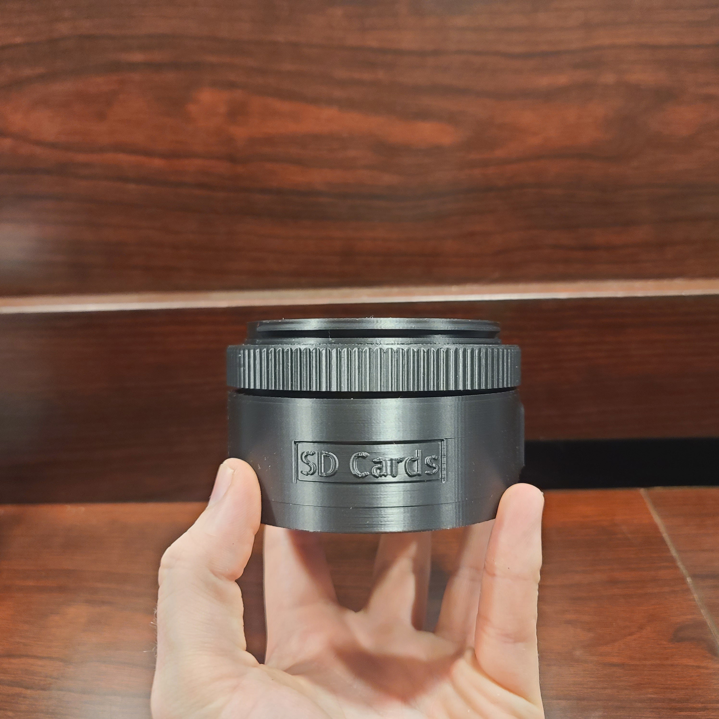 hand holding an sd card holder that spins to display the sd cards. Focal point is the text 'SD Card' There is a wood table in the background with a wooden backing to it.