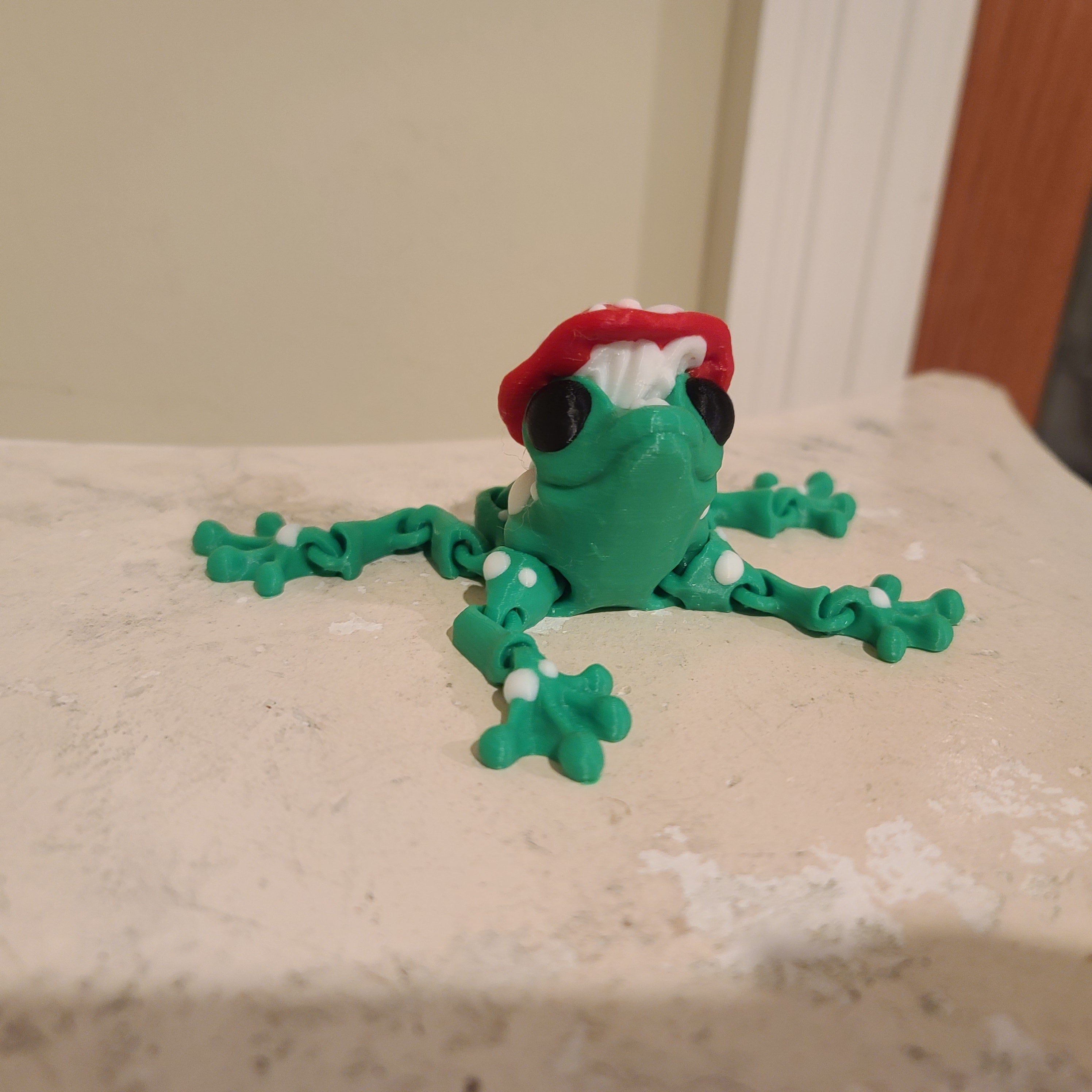 green frog with a red mushroom hat and black eyes and covered with white spots sitting on a stone column in front of some molding.