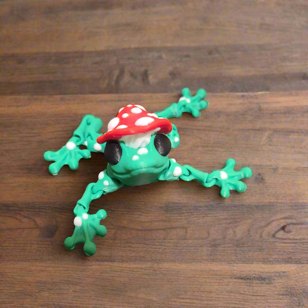 green frog with a red mushroom hat which is covered in white spots and the frog has black eyes and covered with white spots sitting on a wooden table