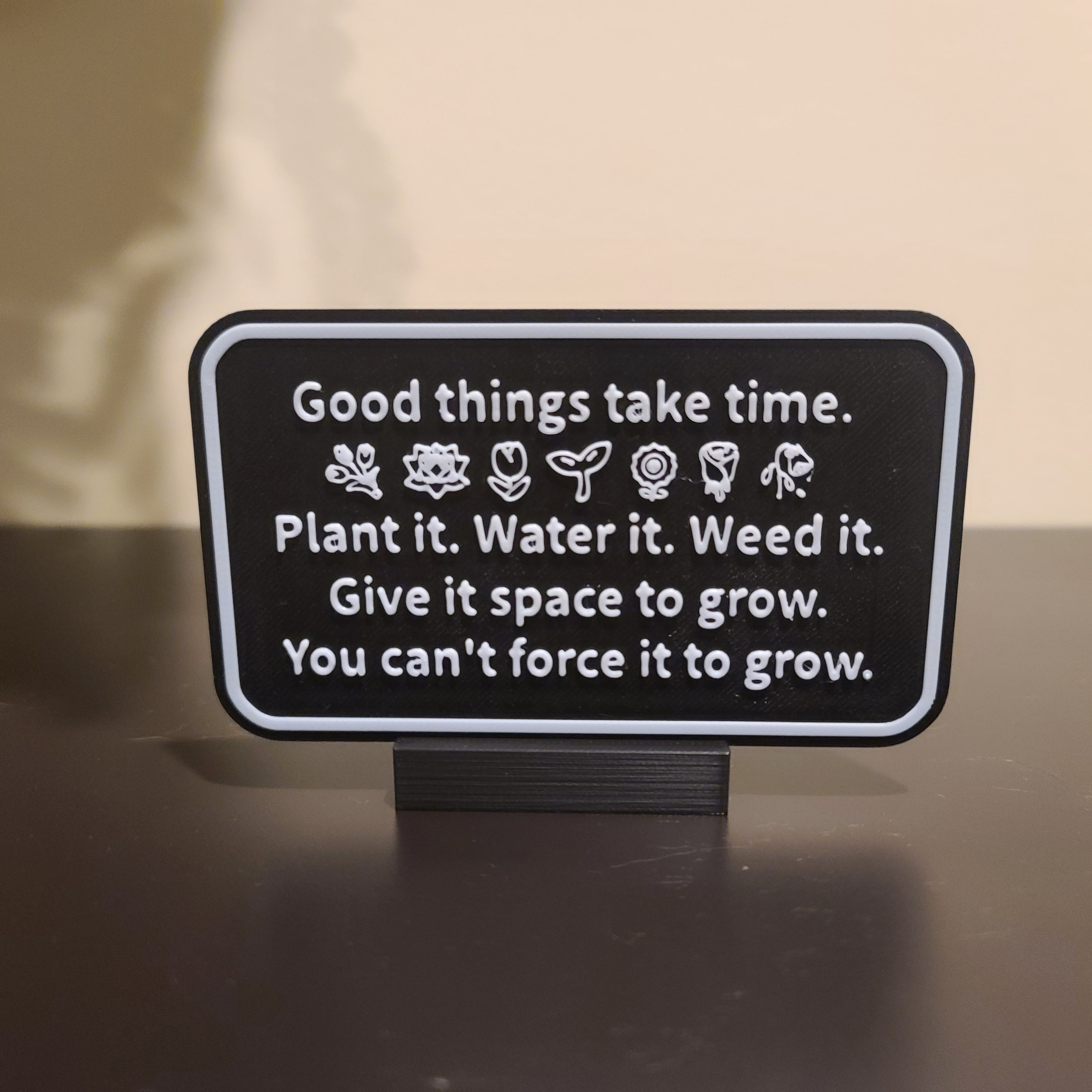 Black table in front of a white wall. There is a sign with the text "Good things take time." Then a row of nature emojis. Followed by the following text: "Plant it. Water it. Weed it. Give it space to grow. You can't force it to grow."