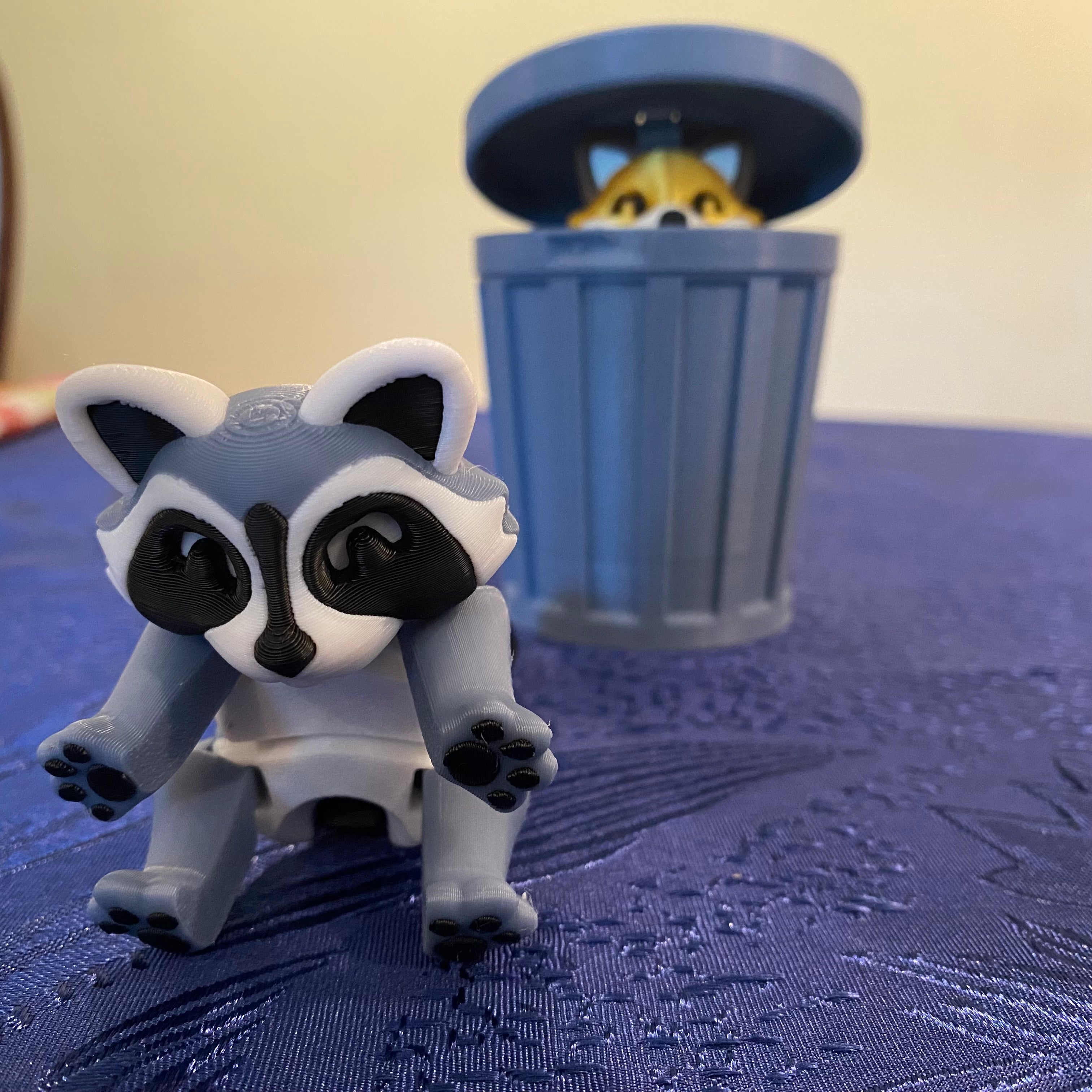 Toy raccoon figure on a blue textured surface with a small trash can in the background.