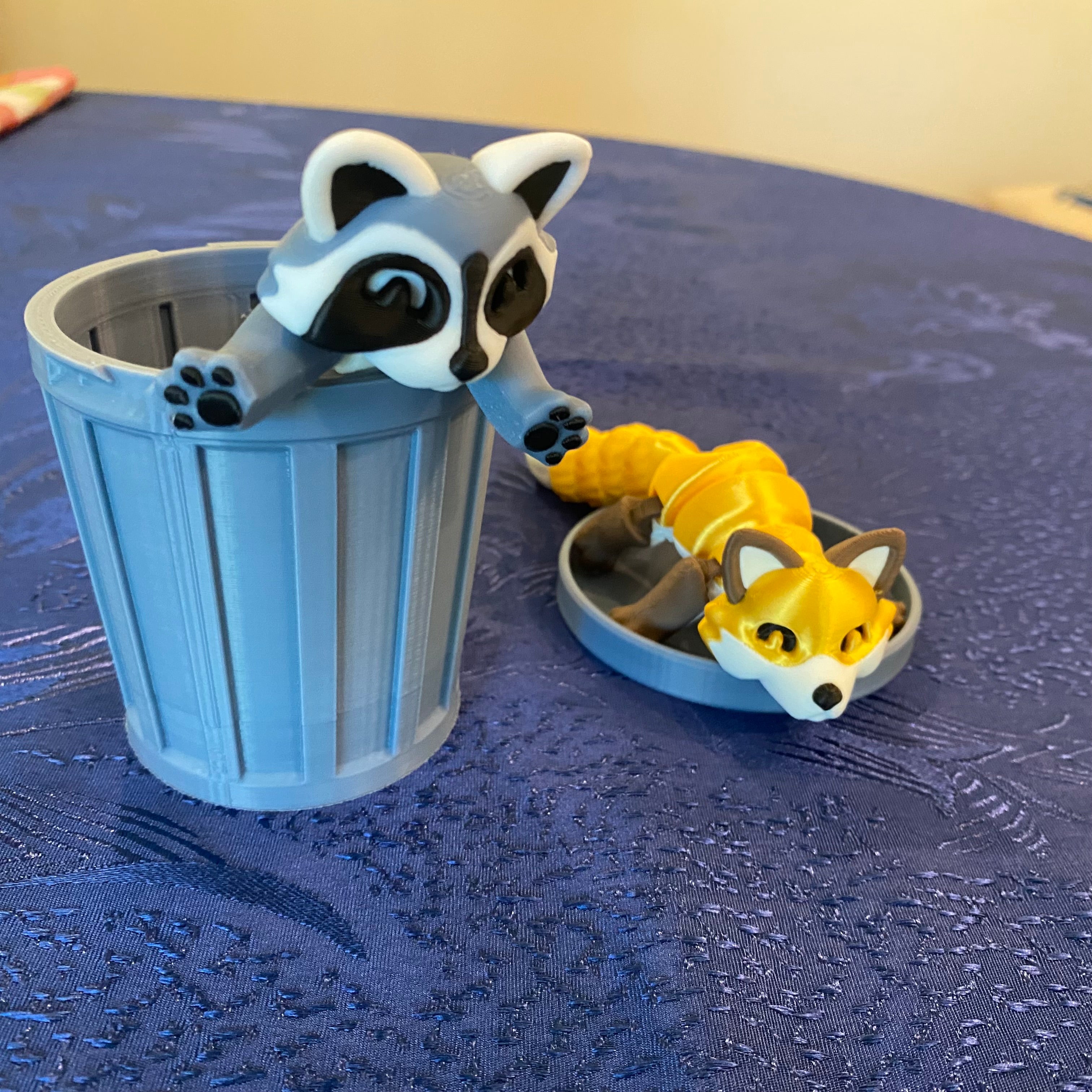 Toy raccoon climbing into a toy trash can on a textured blue surface