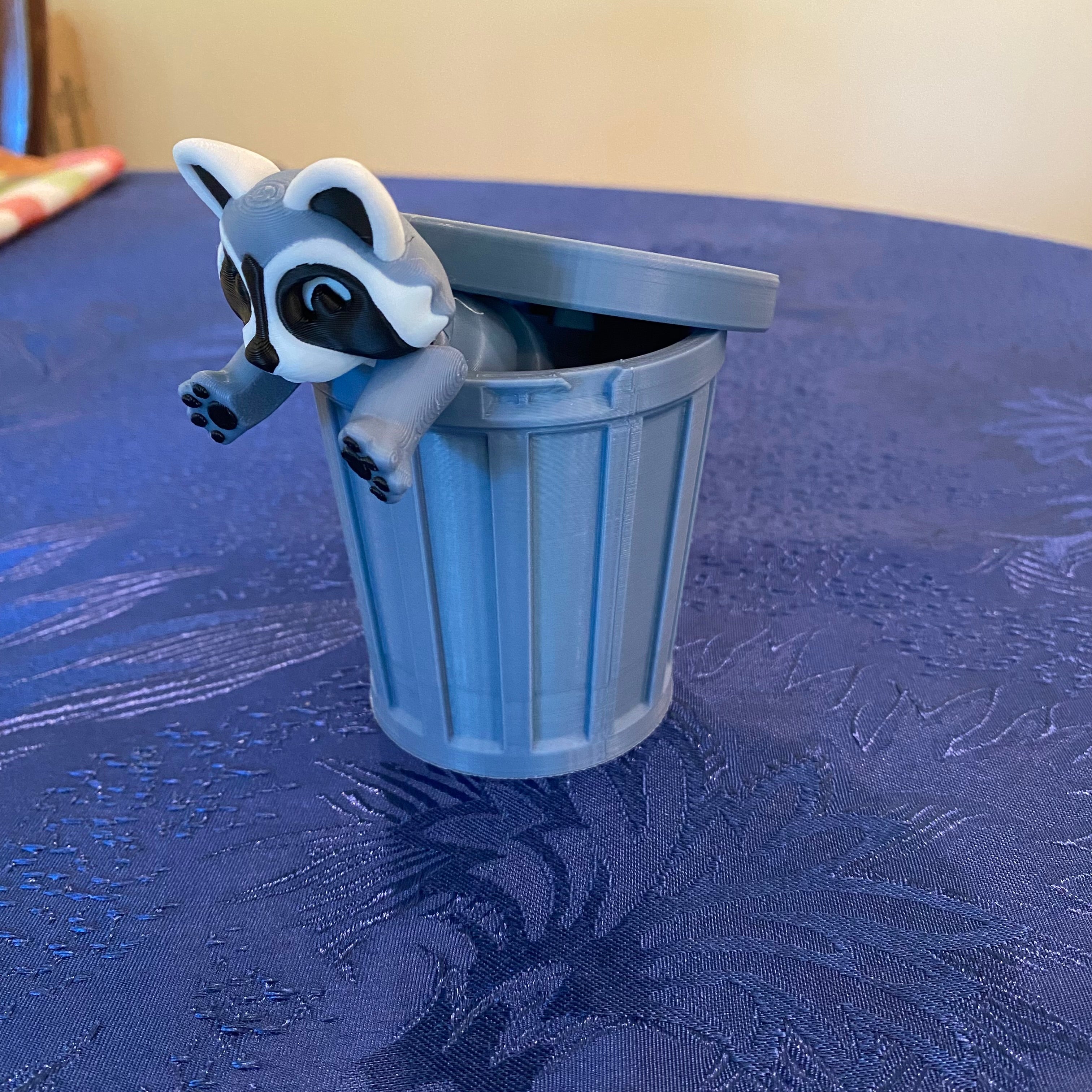 Small figurine of a raccoon climbing out of a blue trash can on a textured surface.