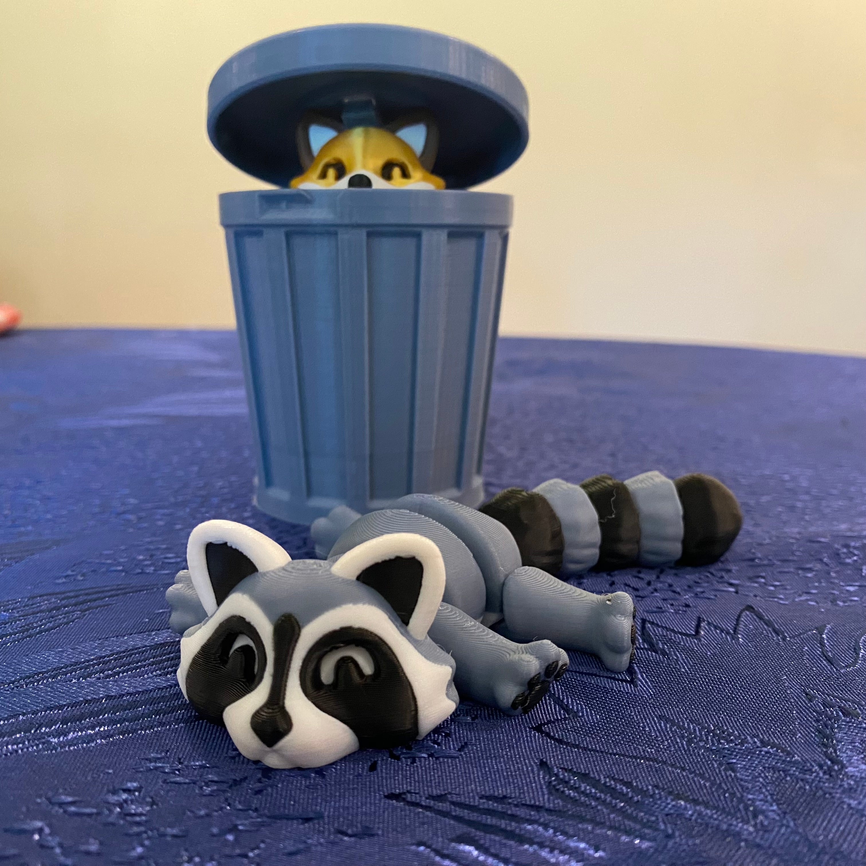 Toy raccoon lying on a blue surface with a toy trash can and fox peeking out.