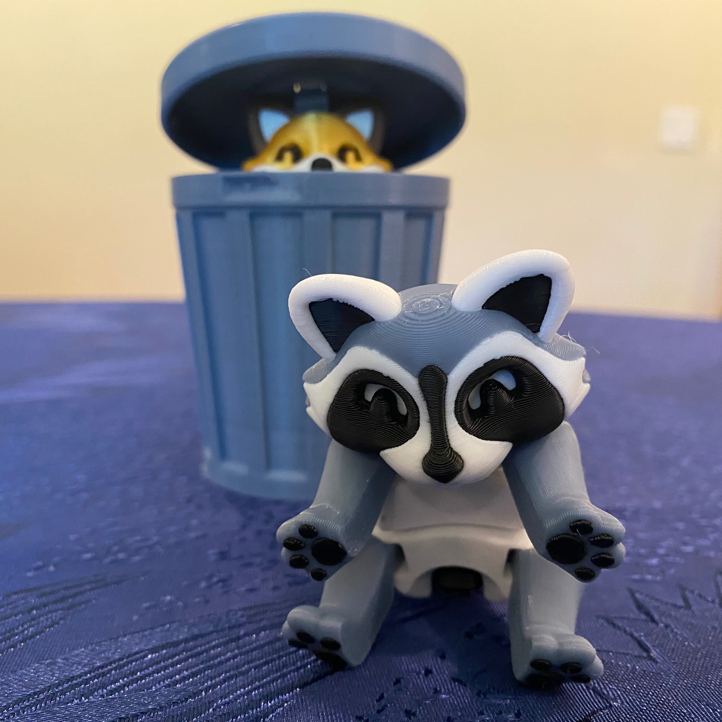 Toy raccoon figure in front of a toy trash can on a blue surface.