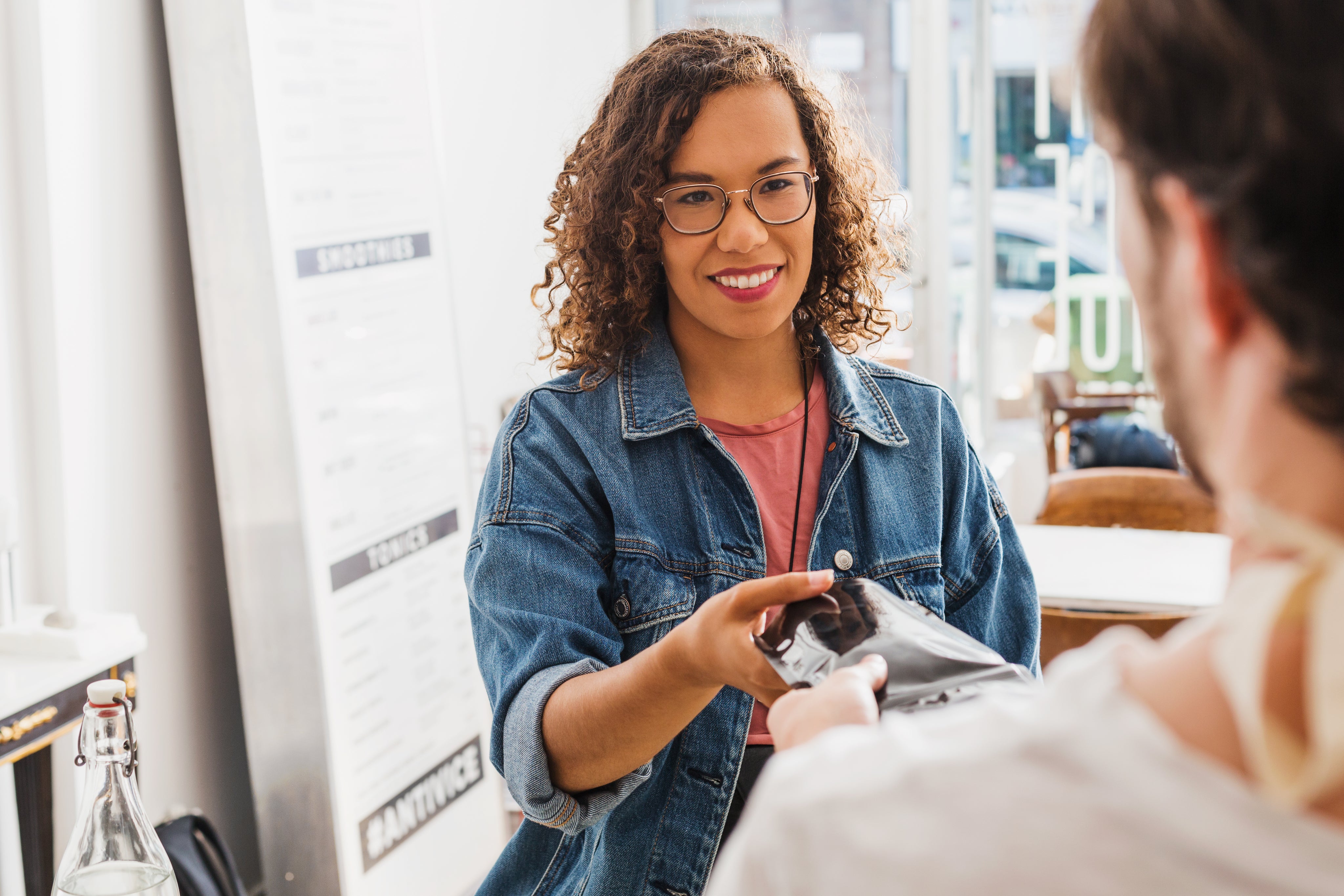 male wearing a white shirt handing a packaged item to a female with curly brown hair wearing a jean jacket.