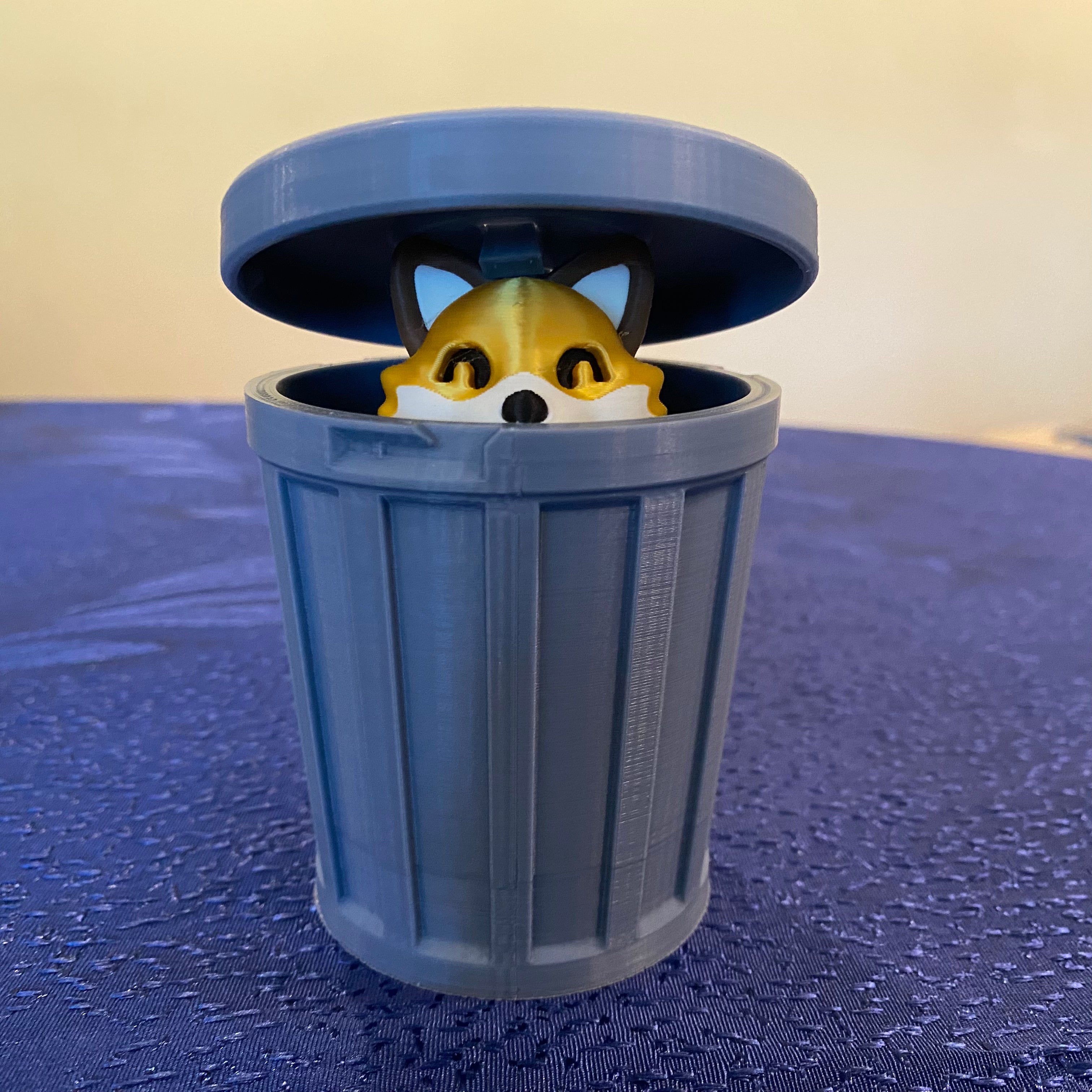 Toy fox peeking out from a blue toy trash can on a blue surface with a beige background