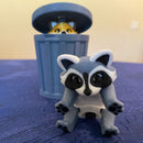 Toy raccoon figure in front of a toy trash can on a blue surface.
